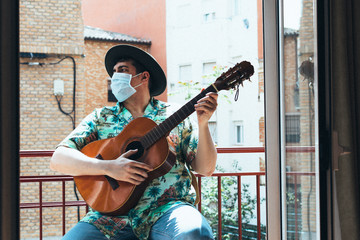 Bohemian musician with mask on his face and hat playing the Spanish guitar from his balcony. Dressed in a Hawaiian shirt and jeans Concept music from home during confinement. Coronavirus pandemic.