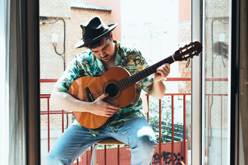 Bohemian musician with mask on his face and hat playing the Spanish guitar from his balcony. Dressed in a Hawaiian shirt and jeans Concept music from home during confinement. Coronavirus pandemic.