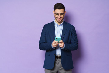 Young business guy isolated on purple background, holding smartphone in hands, looking at screen with interest, laughing at joke sent by friend