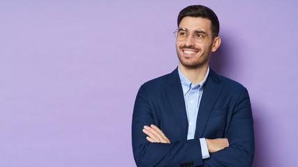 Business guy in eyeglasses dressed in blue formal jacket, smiling happily, standing with arms crossed, looking aside, isolated on purple background, copyspace on left - Powered by Adobe