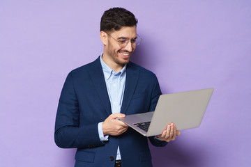 Young business guy dressed in blue suit, wearing glasses, holding laptop in hands, looking at...
