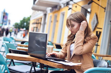 Beautiful young business woman in a beige jacket working with documents and a laptop in a city cafe.	