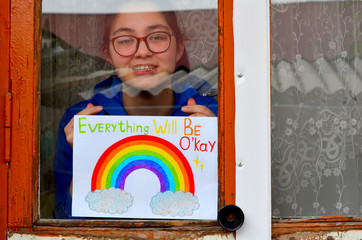 A girl with glasses behind the glass in the window holds a picture of a rainbow symbolizing the imminent end of the quarantine in connection with the coronavirus pandemic.