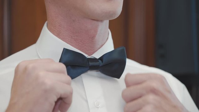 A groom in a white shirt straightens a blue bow tie against a blue sky close-up