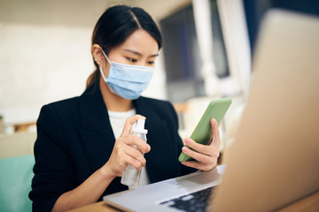 A young Asian woman cleaning and sterilizing her smartphone with alcohol spray at home office.