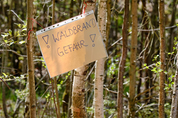 A handwritten sheet of paper in a document wallet hanging between tree trunks and informing about the danger auf Waldbrandgefahr ( english wildfire risk ). Seen in Germany in Bavaria in April.