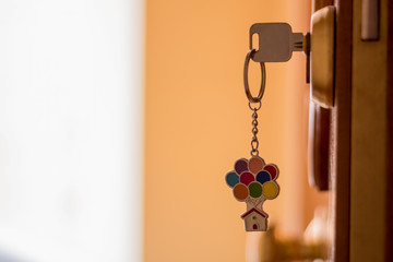 A key with a keychain of a house with colored balloons, on a wooden door with white and yellow background. New normality.