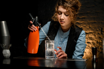 female bartender holds orange siphon and accurate pours water from it