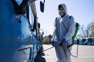 Public transport disinfection. Man in white protective suit with reservoir spraying disinfectant on parked buses. Stop coronavirus or COVID-19.