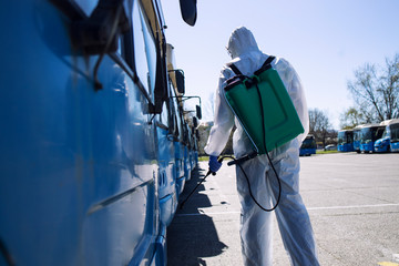 Public transport disinfection. Man in white protective suit with reservoir spraying disinfectant on parked buses. Stop coronavirus or COVID-19.