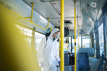 Public transportation healthcare. Man in white protection suit disinfecting and sanitizing handlebars and bus interior to stop spreading highly contagious coronavirus or COVID-19.
