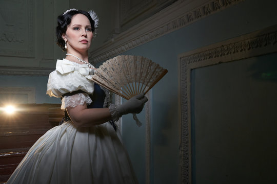 Beautiful girl, in a romantic dress, retro style.With dark hair. Stands against the background of the stage in the theater with a fan, and looks away. Historical reconstruction