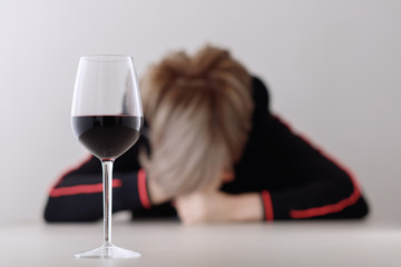 Glass with alcohol on blurred view of a lonely and desperate drunk woman lying on a table. Shallow depth of field. Focus on a wine glass