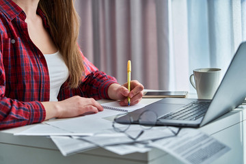 Woman blogger working remotely on laptop and writing down important data information in notebook...