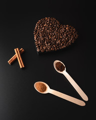 Heart shaped coffee beans, cinnamon sticks and spoons with ground coffee and cinnamon powder on a dark background