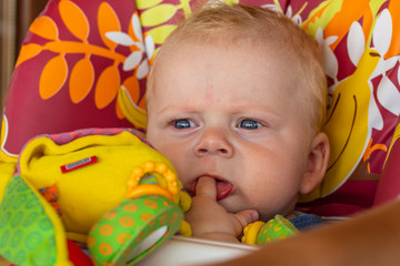 little blond boy sitting in a baby chair at the table with a finger in his mouth while waiting for dinner