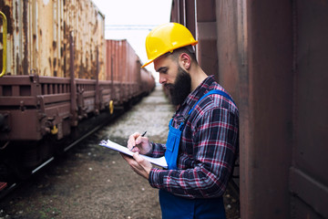 Checking on cargo containers arrived via freight trains. Railway worker writing on his clipboard and keeping track of shipment dispatch. Organizing goods distribution and logistics.