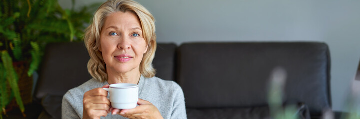 Delicious drink. Cheerful elderly woman sitting on the sofa in her living room and holding a cup of coffee