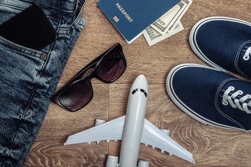 Naklejka premium Travel or tourism concept. Jeans, hundred dollar bills, sneakers, smartphone, passport, sunglasses, airplane on a wooden floor. Top view. Flat lay