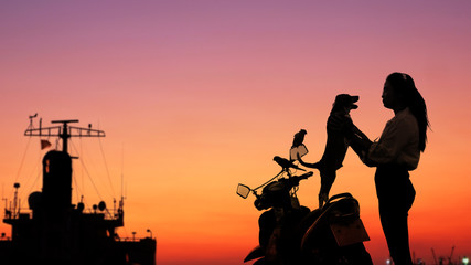Silhouette young woman affectionately playing with her puppy dog and parrots couple on motorcycle at sea viewpoint in harbor with blurred part of ship against colorful sunset sky background  