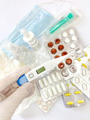 Digital thermometer in hand in glove of a woman on white background with many pills and medicines