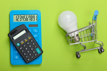Energy saving. Calculators and shopping trolley with led light bulb on green background. Top view