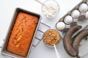 Banana bread in a metal loaf pan is on a kitchen towel. There are eggs, brown sugar, flour and ripe (almost black) bananas near the pan. 