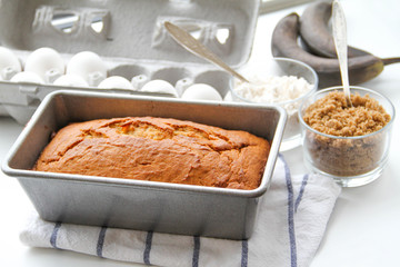 Banana bread in a metal loaf pan is on a kitchen towel. There are eggs, brown sugar, flour and ripe (almost black) bananas near the pan. 
