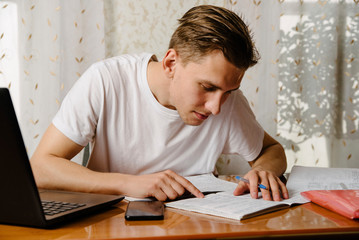 Male young student at home working on a laptop and writing in a notebook.Distance learning
