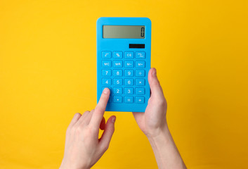 Hand holds blue calculator on yellow background. - obrazy, fototapety, plakaty