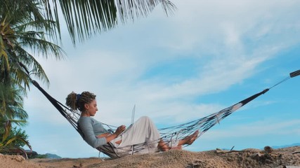 Young woman working on laptop lying in hammock at sand beach of tropical island. Freelance outdoor work concept
