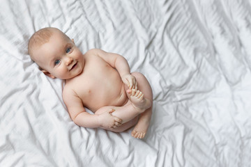 A cute little girl is smiling as she lies on a huge bed with white sheets. Copy space for text. On a white background. Cute little baby lying on bed, cute baby playing with his feet.