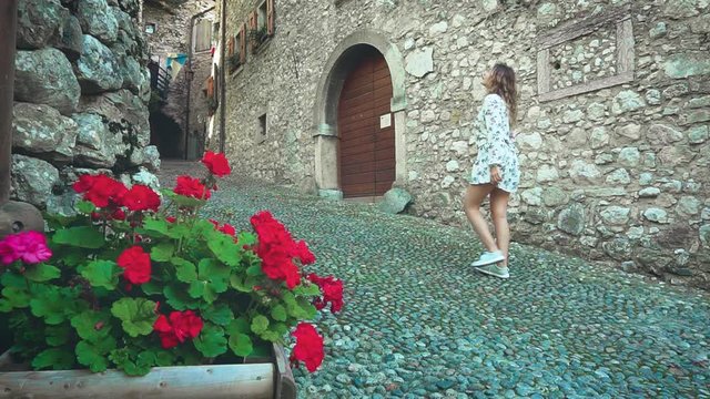 young woman with long blond hair, short sexy white dress walks along stone street of Italian old Medieval Village of Canale Calvola Trentino-Alto Adyghe. Tourist enjoy beautiful architecture flowers