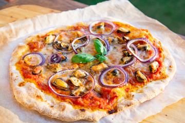 Seafood Italian homemade pizza with mussels, fresh herbs, basil, tomatoes,  red onion and mozzarella on a wooden board. Natural sunny light, close up view.