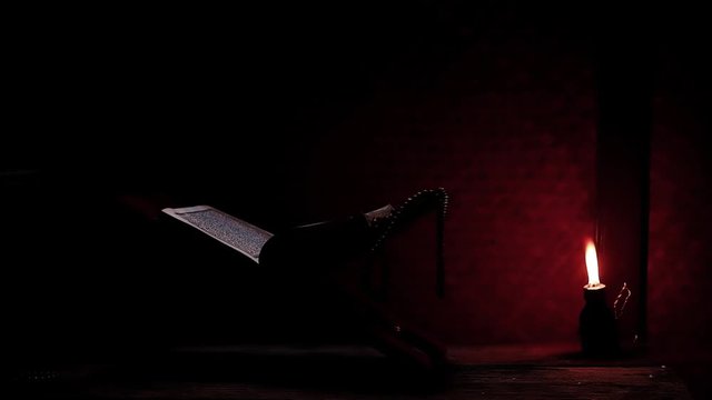 The Quran, the Muslim holy book, with misbaha or the Islamic prayer beads on the rihal or a wooden book stand in a rural room lit by a traditional oil lamp. Ramadan-themed video background.