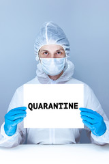 a man in a protective suit, gloves and face mask holds a sheet with the words quarantine in his hands.