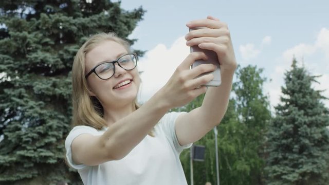 Happy youthful bespectacled female with blue eyes takes selfie and grimaces in park in front of spurce tree and blue sky during warm day. Camera floating around.