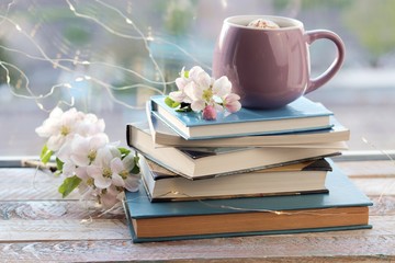 Cup of coffee, a stack of books, apple flowers on a wooden windowsill, home comfort concept   