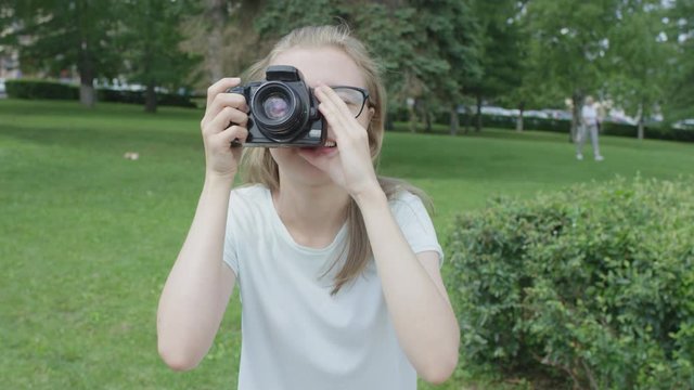 Young bespectacled blonde girl in park holding and adjusting camera pointed stright at viewer looking for a good shot on a summer sunny day.
