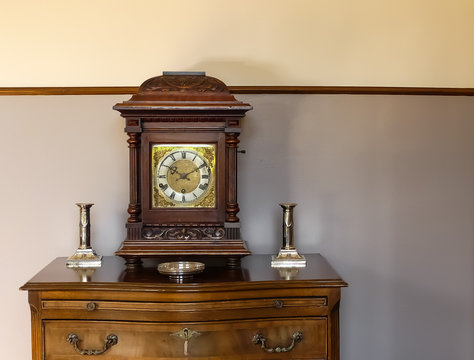 Naturally lit indoor image of a wooden Victorian bracket clock sitting on a mahogany chest of drawers. Flanked by period silver candlesticks. Two tone decoration with dark wood dado rail. England. 