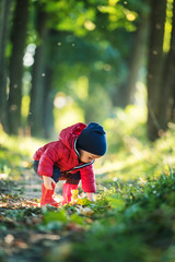 Little toddler boy in red rubber boots and red jacket in spring park. Lush green forest leaves on background
