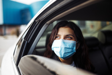 A woman wearing a medical sterile mask in a taxi car on a backseat looking sideway out of open window. Girl passenger waiting in a traffic jam during coronavirus quarantine. Healthcare safety concept.