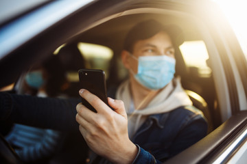 A man wearing a medical mask holds a mobile cell phone in his hand while driving a car. Boy driver checking coronavirus world news in a traffic jam. Isolation and healthcare during quarantine concept.
