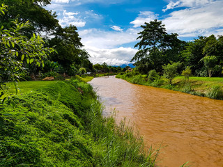 Fototapeta na wymiar Green side next to brown dirt river flow water trees clear blue light sky some clouds pai in chiang mai north thailand