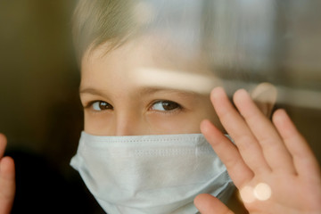Young boy in a medical mask looks out the window. Self-isolation in quarantine, coronavirus, covid 19.