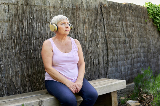 Caucasian Older Woman With Headphones Sitting Outdoors Listening To Music.