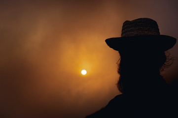 BORȘA, ROMANIA – JULY 07 2015: Man with beard and long hair wearing a hat, silhouette in mountain setting with the sunset behind