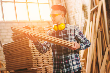 Young skilled carpenter in protective eyewear and headphones working with plywood material at sawmill factory warehouse. Professional male cabinet maker at woodworking workshop. Man artisan, DIY - obrazy, fototapety, plakaty