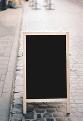 A black wooden billboard stands on a pavers stones. Outdoor advertising concept for cafe and shop. Copy space, mockup.