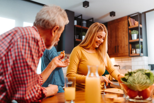 Blonde Woman Is Chopping Vegetables For Salad While Her Father Standing Next To Her Eating Pretzels And Drink Freshly Squeezed Juice.